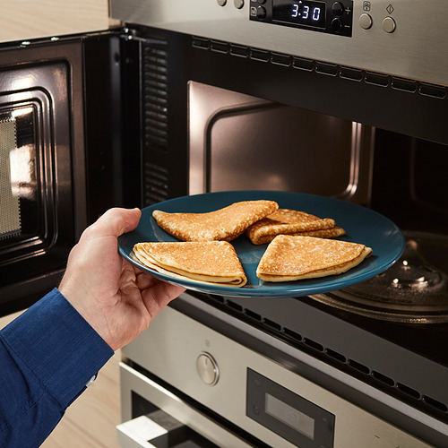 Una persona sosteniendo un plato azul con panqueques dorados cerca de un horno de microondas.
