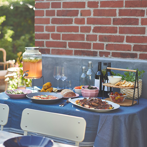 Mesa de exterior con bandeja FULLSPÄCKAD azul, asa de bambú, que sirve varios platos y bebidas contra la pared de ladrillo rojo.