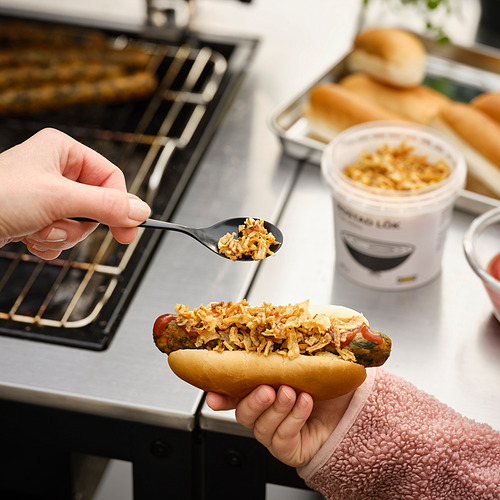 Una persona prepara un perrito caliente, agregando aderezos de cebolla de un recipiente etiquetado como ROSTAD LÖK. La configuración de la cocina muestra una estufa de gas y otros artículos de cocina.