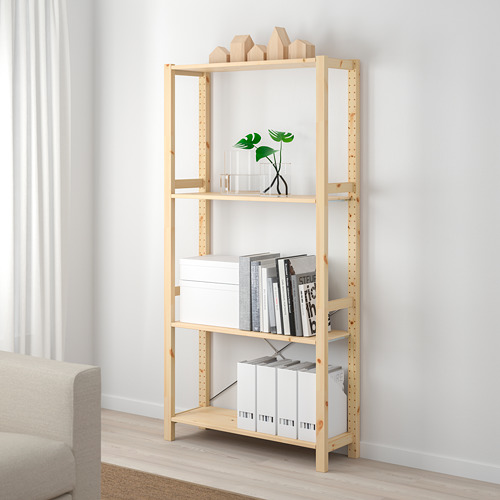 Una estantería de pino marrón claro IVAR. Se alza junto a un sofá beige, con libros, cajas y una planta.