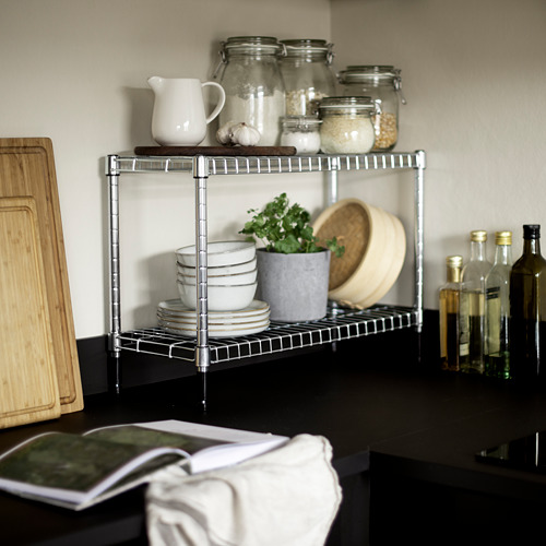 Encimera de cocina con estante de almacenamiento de plata OMAR que contiene frascos, tazones y una planta. Tabla de cortar de madera y botellas cerca.