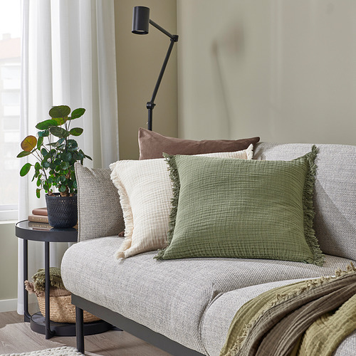 Acogedora sala de estar con sofá gris claro, cojines VALLKRASSING verdes, mesa auxiliar con plantas y libros.
