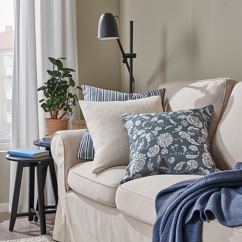 Cojines IDALINNEA sobre sillón beige, floral blanco y azul, junto a una mesita negra con plantas.