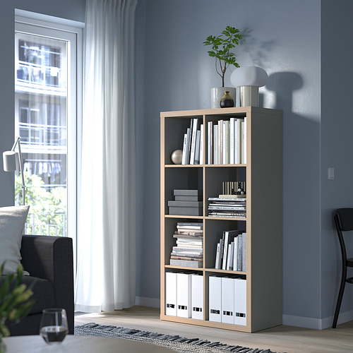 Estantería KALLAX con libros y decoración, junto a un sofá gris y una silla negra, contra una pared azul.