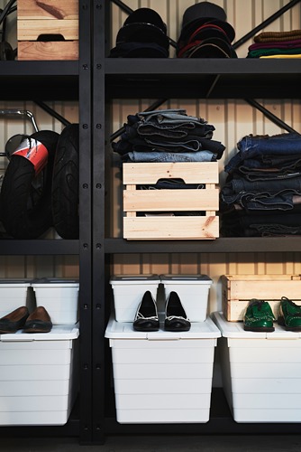 Un sistema de almacenamiento de metal negro, BROR, con estantes organizados. Contiene contenedores blancos, ropa doblada y cajas de madera.