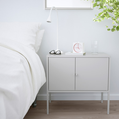 Un armario de almacenaje blanco de IKEA LIXHULT junto a una cama. Es compacto, con dos puertas, y soporta una lámpara, un reloj, gafas y un jarrón.