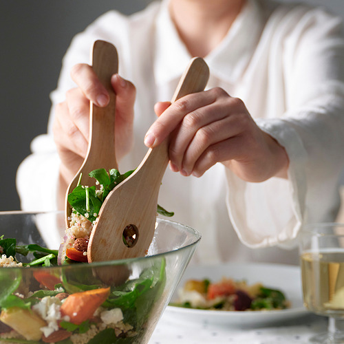 Una persona sostiene pinzas de madera agarrando una ensalada verde de un tazón transparente. Un paño blanco y un vaso están cerca.