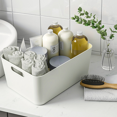 Caja organizadora blanca en la encimera del baño que almacena toallas cuidadosamente dobladas, botellas de champú y una crema facial, con asas para facilitar su transporte.