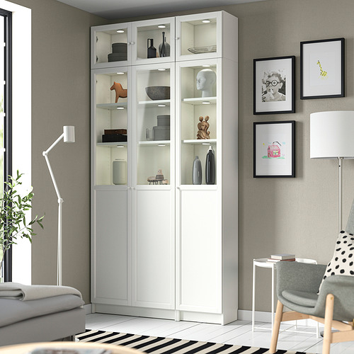 Librería blanca BILLY con puertas de vidrio y madera, artículos decorativos, arte cerca de la ventana y la pared, sofá gris, silla de lunares.