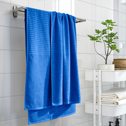 La toalla VÅGSJÖN azul brillante cuelga en un estante plateado sobre estanterías blancas con toallas dobladas y planta verde, pared de azulejos blancos en contraste.