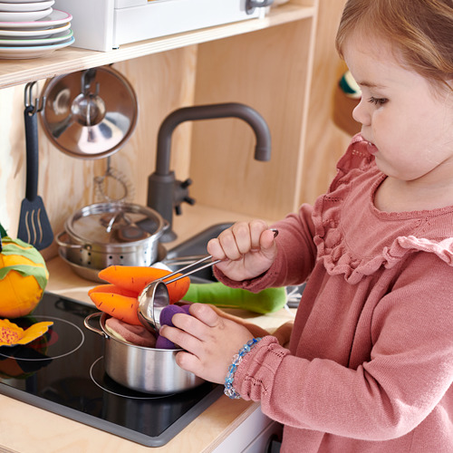 Niña con suéter rosa usando una pequeña cuchara de plata para cocinar verduras coloridas en la mini cocina.