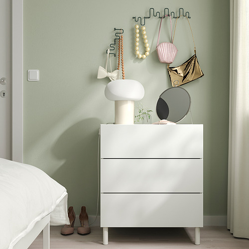 Dormitorio moderno con tocador blanco, lámpara, espejo, plantas y ganchos verdes que sostienen bolsos. Las cortinas de luz enmarcan la escena.