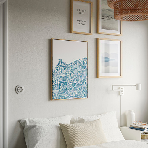 Pared con obras de arte y cama. Centrado: póster BLODFLÄDER de onda azul en marco de madera clara. Ropa de cama blanca debajo. Estética serena y moderna.
