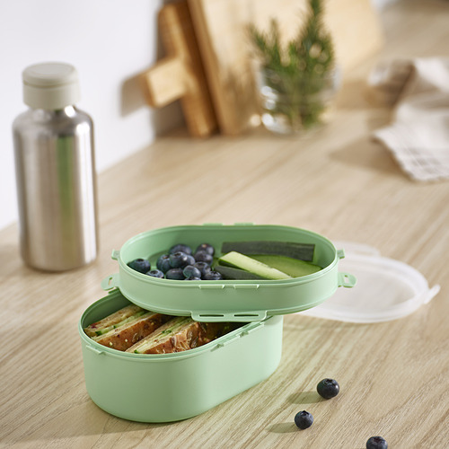Fiambrera ovalada de plástico verde menta UTBJUDA, tamaño similar a la botella de agua, dividida en dos niveles para alimentos, colocada sobre mesa de madera clara.