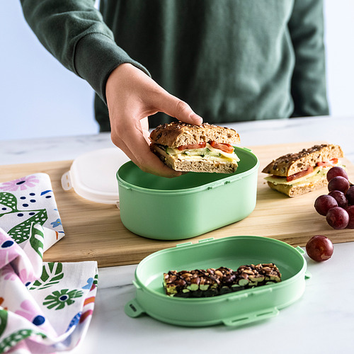 La mano coloca el sándwich en una lonchera verde de dos niveles, que separa la comida en una tabla de madera con uvas y una servilleta estampada.