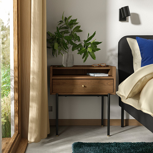 Escena de dormitorio con mesita de noche RÅDMANSÖ de mediados de siglo en cálido nogal con patas de metal negro, cortinas color crema y una almohada azul sobre una cama.