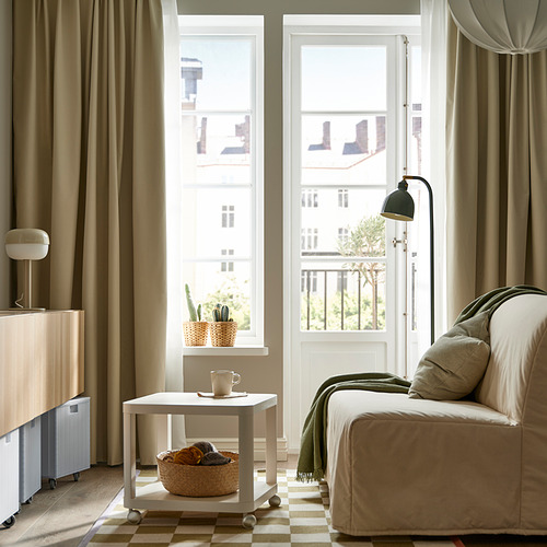 Dormitorio moderno con lámpara de color oliva, sofá beige, mesa auxiliar con ruedas y gran ventanal.
