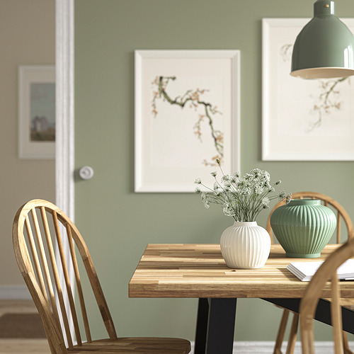 Acogedora zona de comedor con mesa de madera, jarrón PELARBJÖRK con flores, bajo lámpara moderna, pared verde salvia con cuadros.