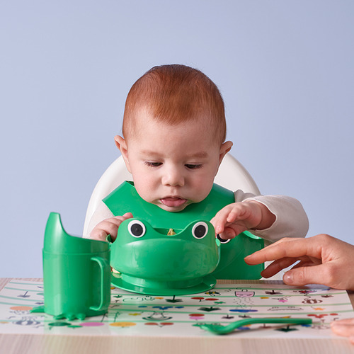 Bebé en silla alta con vaso de rana verde. Un adulto asiste en coloridos manteles individuales.