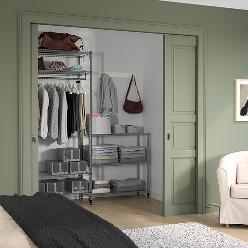 Estantería verde OMAR con estantes metálicos ajustables, ropa ordenada y bolsos en una acogedora sala verde.
