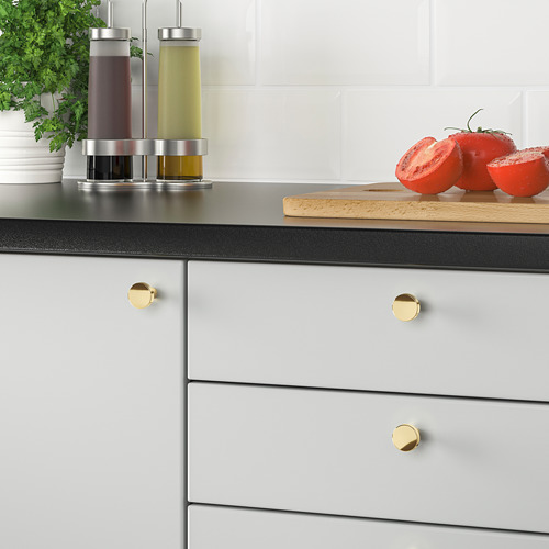 Cocina moderna con pomos de latón BAGGANÄS. La encimera negra contiene tomates y hierbas.