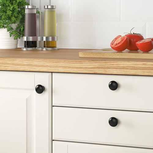 Encimera de cocina de madera con cajones blancos con pomos negros ENERYDA. Cerca de allí, hay botellas de colores, tomates frescos y una planta.