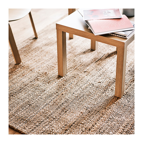 Alfombra de yute beige natural, rectangular, textura gruesa, sobre suelo de madera, debajo de una mesa de madera clara con patas visibles, colocadas cerca de las sillas.