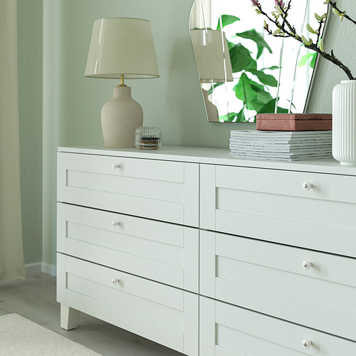 Cómoda blanca con seis cajones. Lámpara decorativa, espejo, pila de libros y jarrón. Los pomos son de porcelana blanca.