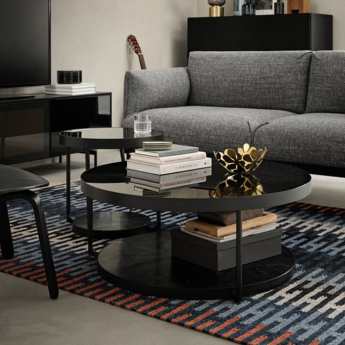 Sala de estar moderna con sofá gris, mesa auxiliar negra y alfombra colorida. La mesa contiene libros, vidrio y arte decorativo.