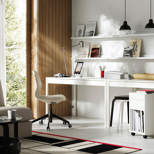 Moderna oficina en casa con escritorio, silla y estantes blancos. El cajón y los estantes contienen libros y decoración. Luz natural y una alfombra roja/negra.