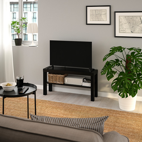 Sala de estar moderna con banco de televisión LACK bLACK, TV de pantalla plana, sofá gris, mesa auxiliar redonda con cuenco y bebida, planta grande, lámpara y mapas de pared.