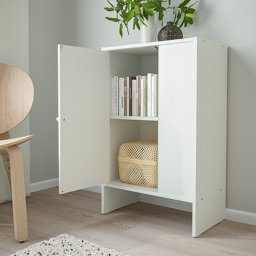 Pequeño armario blanco BAGGEBO con puertas, libros de almacenamiento y una cesta, montado contra una pared de color verde claro.