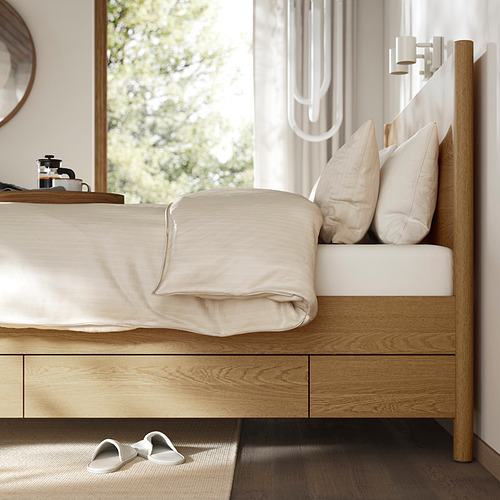 Cama moderna de madera con cajones, ropa de cama color crema y zapatillas debajo.