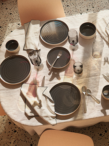 Elegante mesa OMBONAD con cuencos de cerámica oscura, cristalería transparente y servilletas beige sobre una mesa de madera para un diseño rústico minimalista.