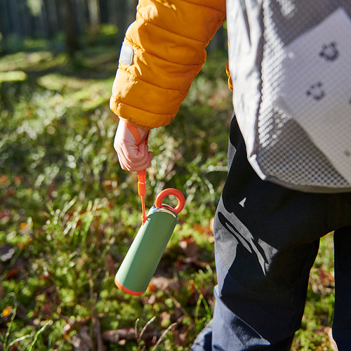 La mano en la chaqueta amarilla sostiene una botella de agua verde-roja con luz naranja.