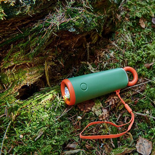 Linterna verde con detalles en naranja y cordón, descansando sobre el suelo musgoso del bosque. Materiales de calidad para mayor durabilidad.