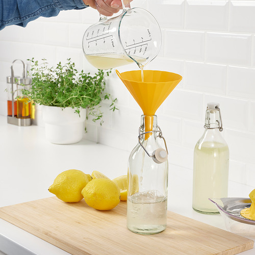 La persona vierte líquido de la taza medidora a través del embudo amarillo en una botella de vidrio. Limones, otra botella y hierbas cerca.