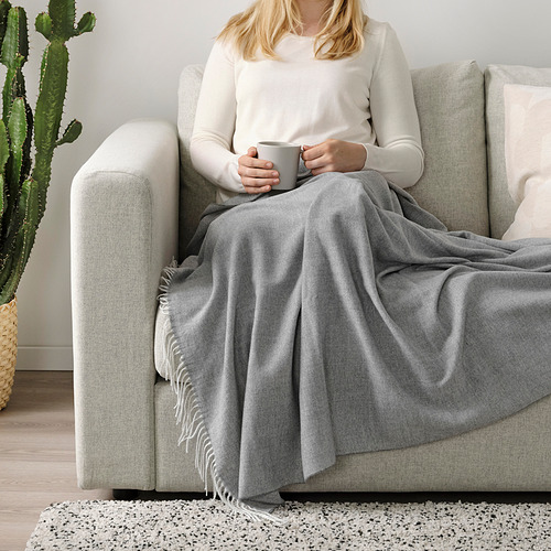 Mujer sentada en un sofá gris, envuelta en una manta de flecos grises, sosteniendo una taza.