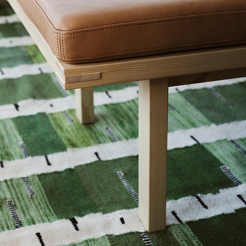 Un taburete de cuero beige con patas de madera clara se asienta sobre una alfombra verde tejida a mano con un patrón de abedul, enfatizando la comodidad y el estilo.