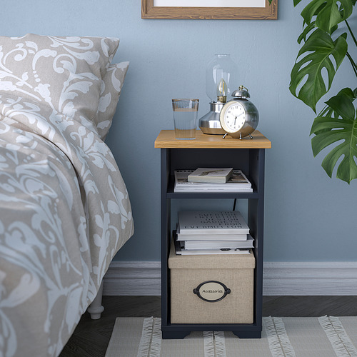Mesa auxiliar SKRUVBY con tapa de madera, estantes abiertos y caja tejida, al lado de la cama con libros, reloj, lámpara.