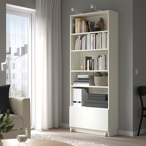Una estantería blanca BILLY con cajones en una sala de estar. Es alta, rodeada de otros muebles, con estanterías llenas de libros y decoración.
