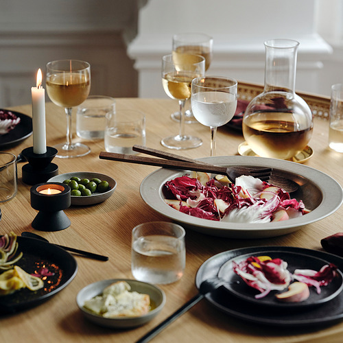 Mesa de madera con platos de gres beige, ensalada de colores, velas, copas de vino y jarra con vino blanco. Centro de mesa con utensilios de madera.