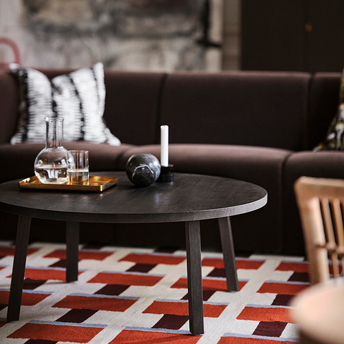 Mesa de centro STOCKHOLM 2025 de color marrón oscuro con tapa de roble sobre alfombra de colores, sosteniendo una jarra, vasos y una vela. Más grande que la silla de madera cercana.