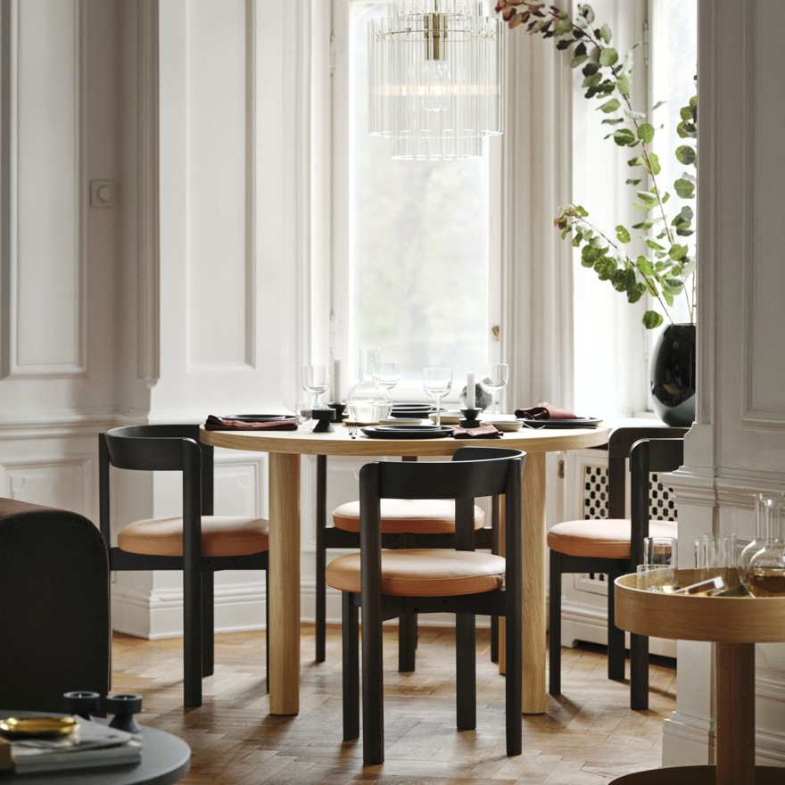 Elegante comedor con mesa de madera clara y sillas negras, candelabro moderno, planta y vajilla.