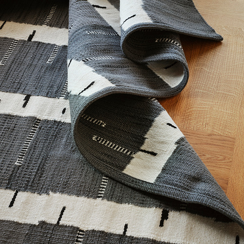 Alfombra de lana tejida a mano en blanco y negro con patrón de abedul parcialmente enrollada sobre suelo de madera. Artesanía de alta calidad visible en la textura detallada.