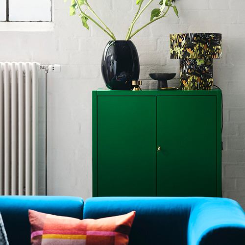 Sofá azul brillante con almohadas de colores, gabinete verde, jarrón oscuro con vegetación, lámpara estampada, radiador blanco a la izquierda, pared de ladrillo gris claro.