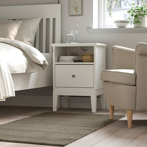 Dormitorio con mesita de noche TIDTABELL, cama blanca, sillón, alfombra gris, acogedor diseño minimalista.