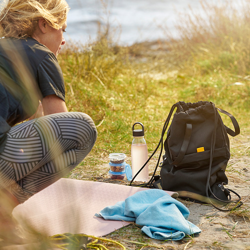 Mujer sentada al aire libre con una botella de agua reutilizable, una mochila y algunos bocadillos. La botella tiene una correa para facilitar su transporte.