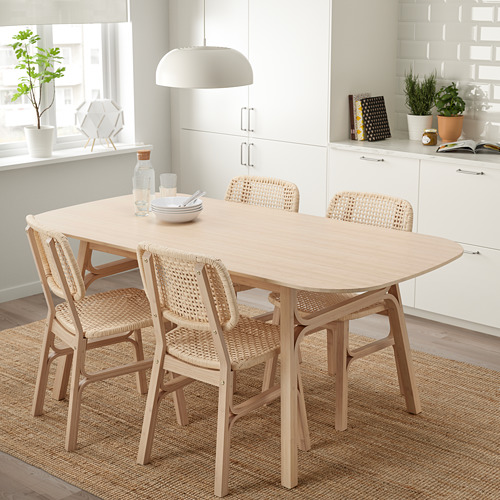 Juego de comedor de bambú con mesa lisa, patas en ángulo y sillas tejidas sobre alfombra de yute, cocina blanca, ventana grande.