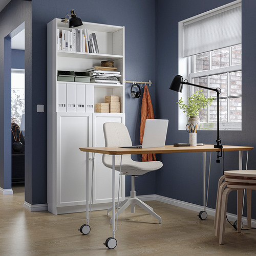 Moderna oficina en casa con estantería blanca BILLY, escritorio de madera clara, silla gris, lámpara negra y ventana.
